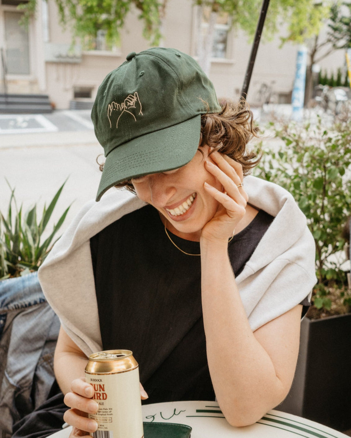 Person sitting at an outdoor cafe table with a can of beer and a glass, wearing a green cap.