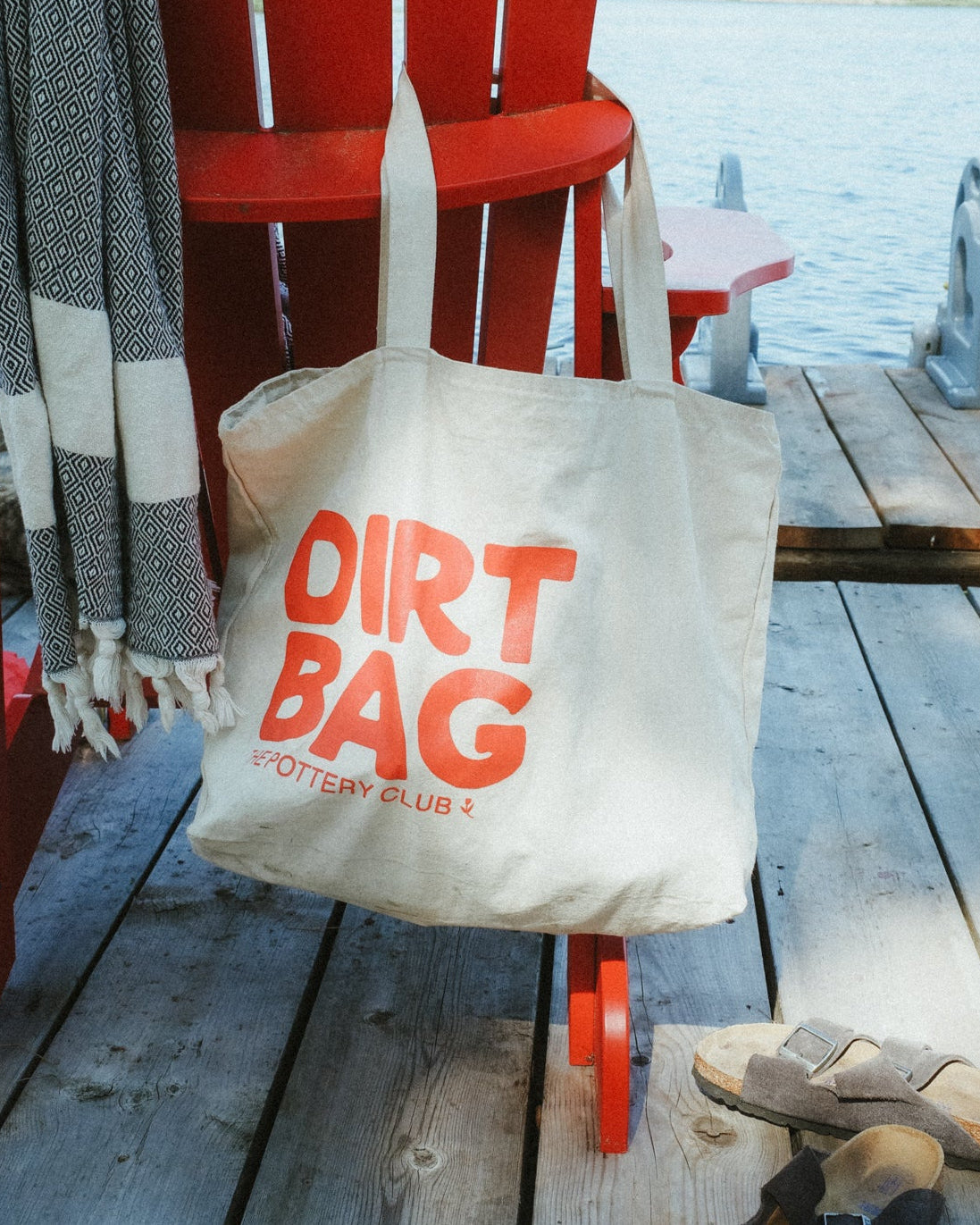 Red Adirondack chair with a 'Dirt Bag' tote bag on a wooden deck by a lake.