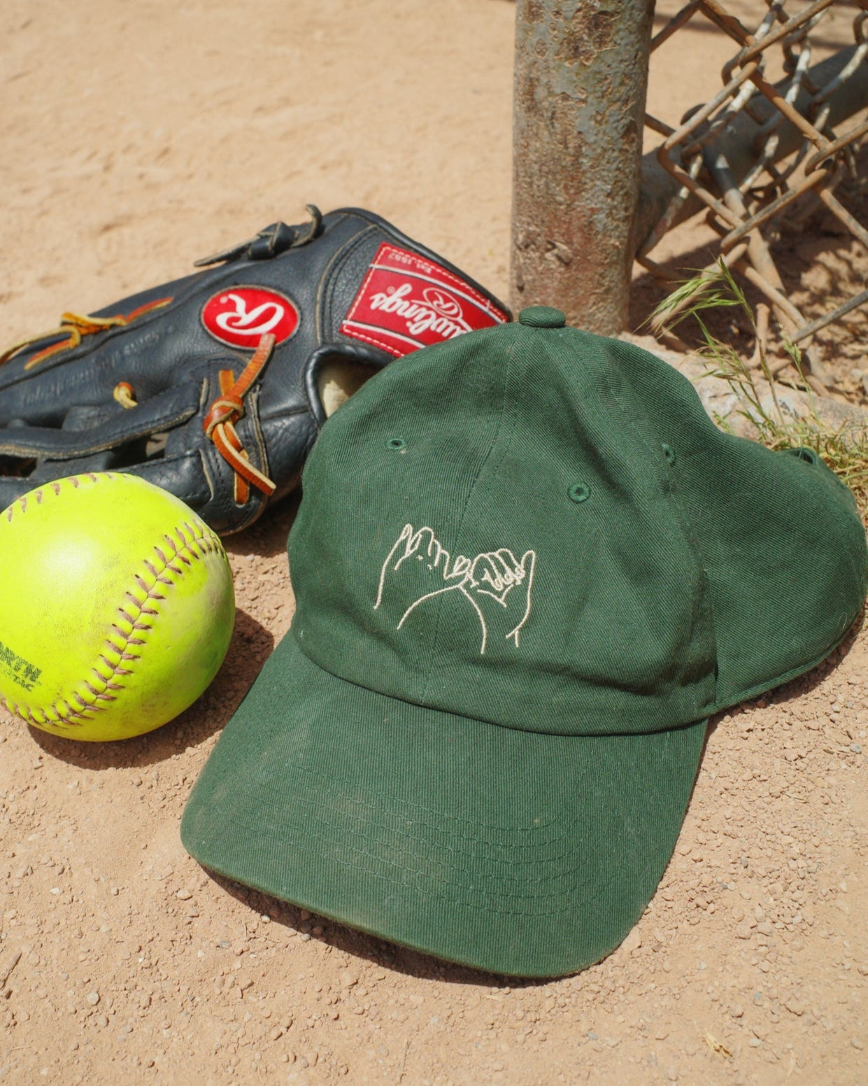 Green cap, black baseball glove, and yellow softball on a dirt surface with a chain-link fence in the background.