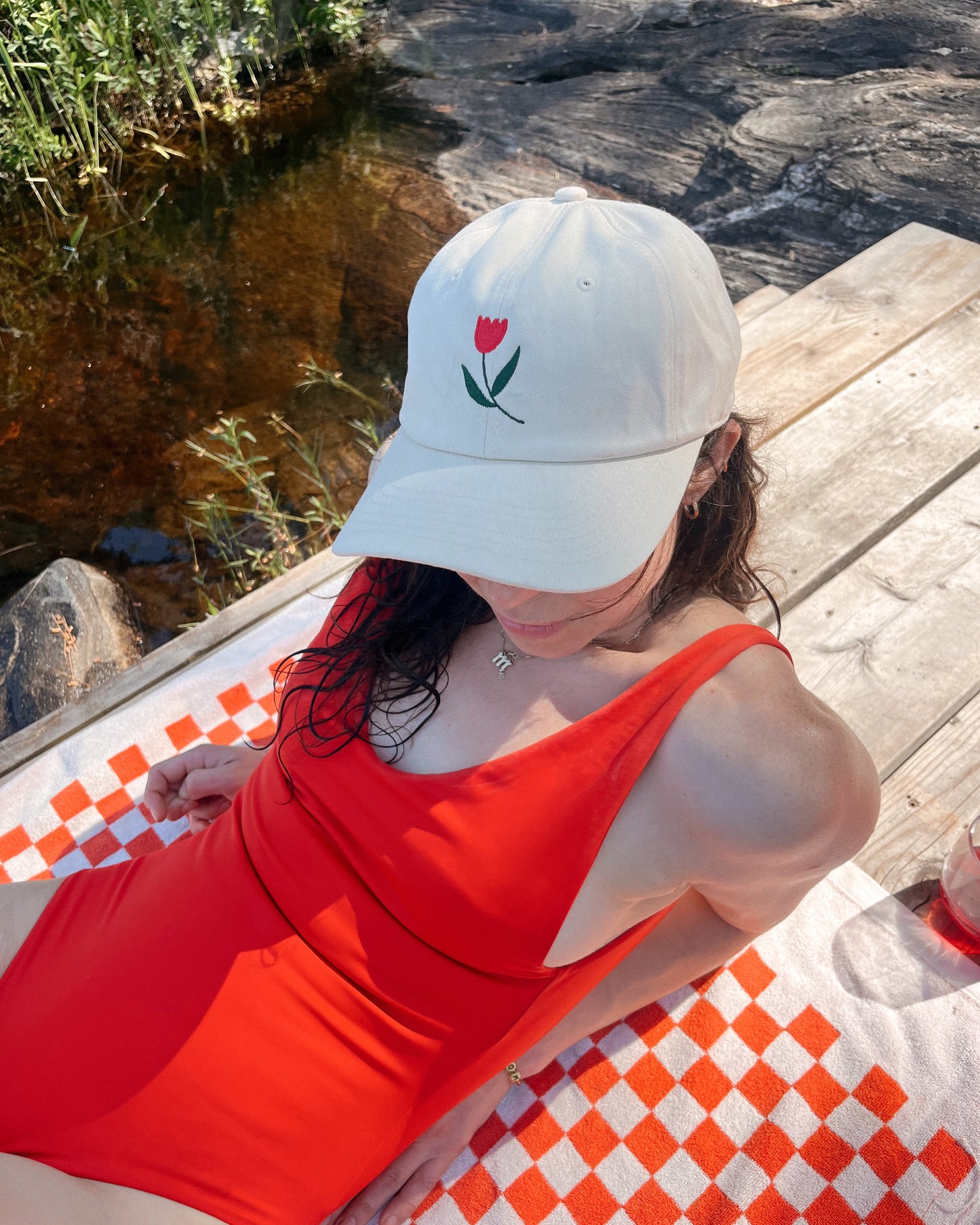 Person wearing a white cap with a red tulip design and a red swimsuit, sitting on a checkered surface by a natural setting.