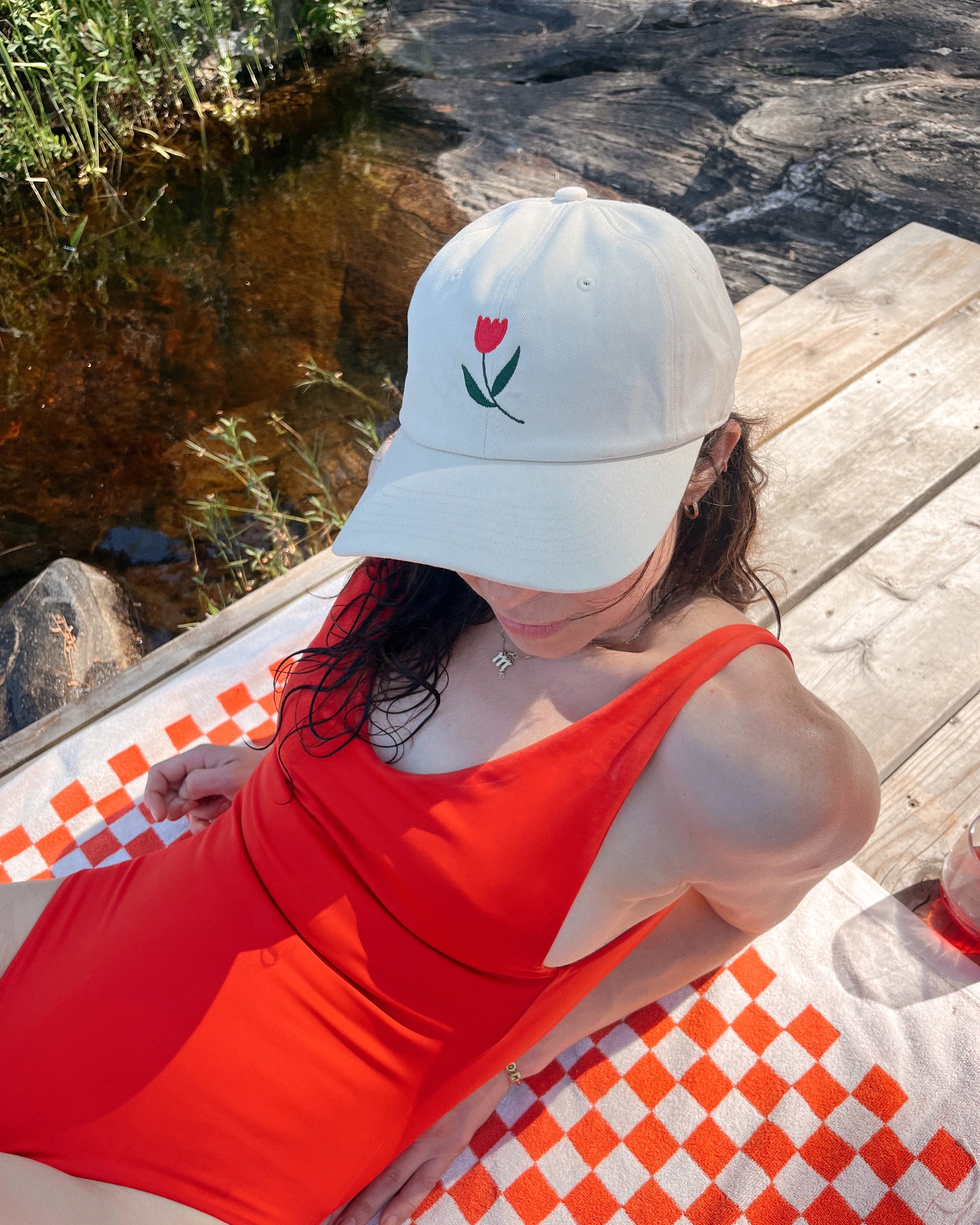 Person wearing a white cap with a red tulip design and a red swimsuit, sitting on a checkered surface by a natural setting.